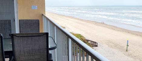 A balcony with a view of a sandy beach and the ocean. There is a chair and a table on the balcony.