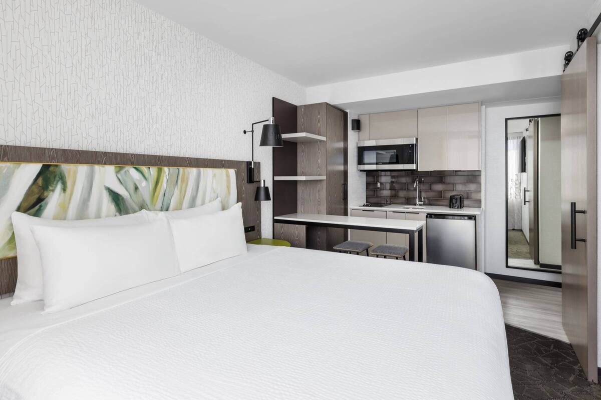 A modern hotel room with a large bed, a desk, and a kitchenette.