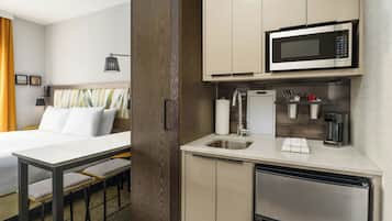 Una habitación de hotel compacta con una cama, una pequeña mesa de comedor con sillas, una kitchenette con microondas, cafetera y lavamanos.