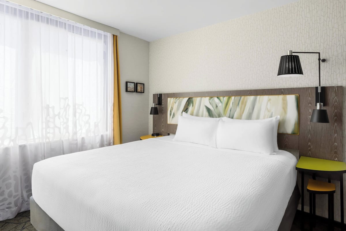 A large bed with white bedding, a wooden headboard, two bedside lamps, a small table with a yellow top, and a window with sheer curtains.