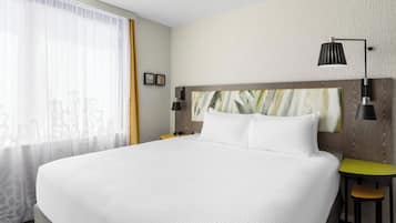 A large bed with white bedding, a wooden headboard, two bedside lamps, a small table with a yellow top, and a window with sheer curtains.