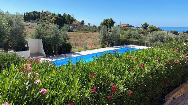 A swimming pool surrounded by lush greenery and pink flowers, with a view of the sea and hills in the background. - Apartment - Sleeps 3 & Swimming Pool (Realmonte)