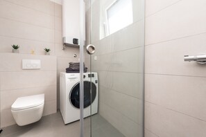 A compact bathroom with a washing machine, toilet, and a glass shower enclosure. - Apartment Birgir in Sukošan (Sukosan)