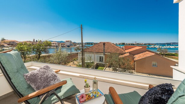 A balcony with a view of a coastal town, a table with a drink, and a chair with a cushion. - Apartment Birgir in Sukošan (Sukosan)