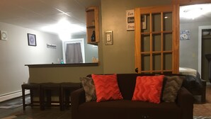 Ein Wohnzimmer mit einem dunkelbraunen Sofa, zwei roten Kissen, einem gemusterten Teppich, einem Holztisch und einer Küche im Hintergrund.