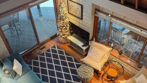 A cozy living room with a fireplace, a Christmas tree, and a large window overlooking a snowy deck. - Cottage on the Bluff - NEW Stairs to the Beach, Hot Tub & Unbeatable Sunsets! (South Haven)
