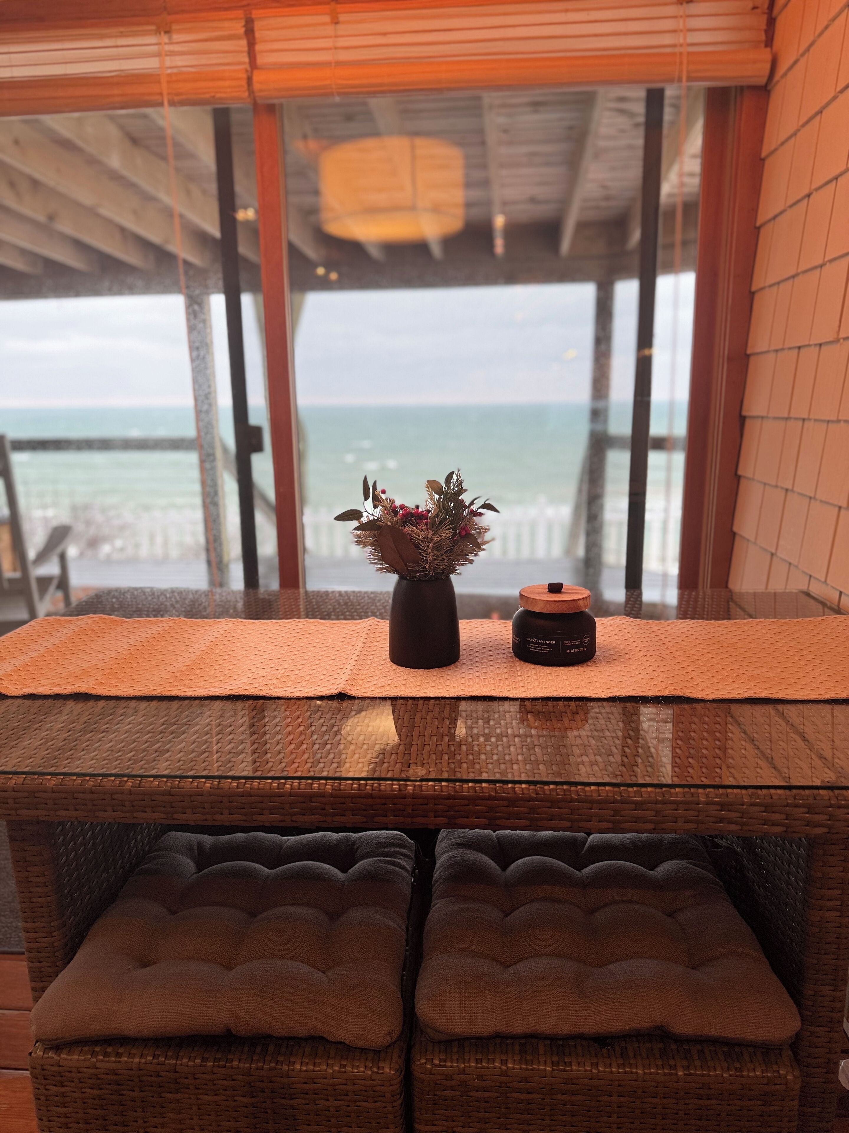 Une chaise en osier avec un coussin, une petite plante en pot et un bocal posés sur une table près d’une fenêtre donnant sur la mer.