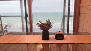 A wicker chair with a cushion, a small potted plant, and a jar on a table by a window overlooking the sea. - Cottage on the Bluff - NEW Stairs to the Beach, Hot Tub & Unbeatable Sunsets! (South Haven)
