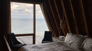 A room with a bed, a chair, and a view of the sea through a large window. - Cottage on the Bluff - NEW Stairs to the Beach, Hot Tub & Unbeatable Sunsets! (South Haven)