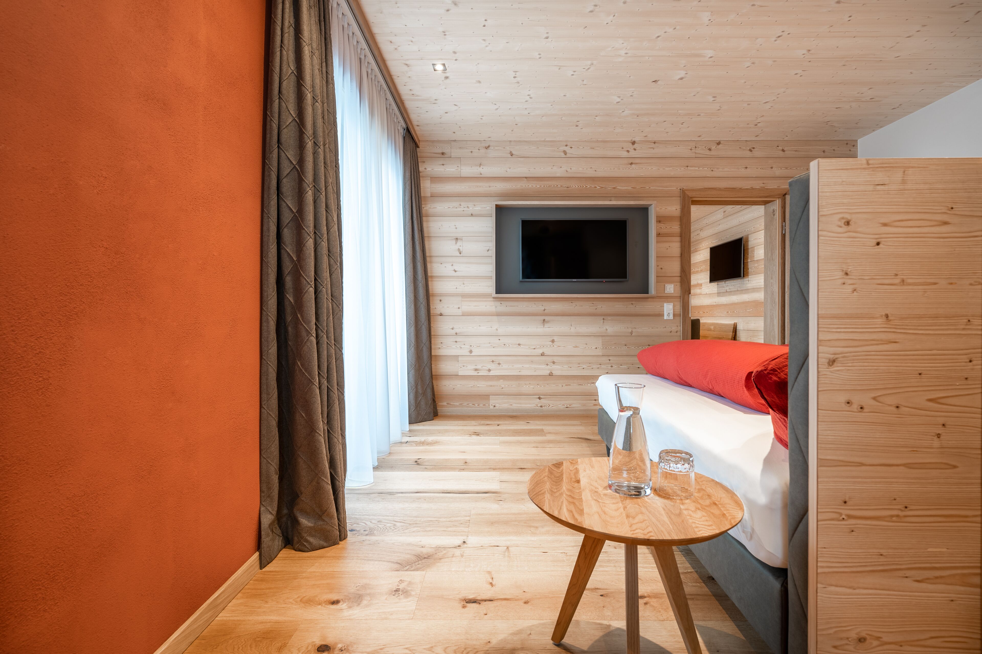 Een kamer met houten lambrisering, een bed, een aan de muur bevestigde televisie en een klein houten tafeltje met een glas erop.