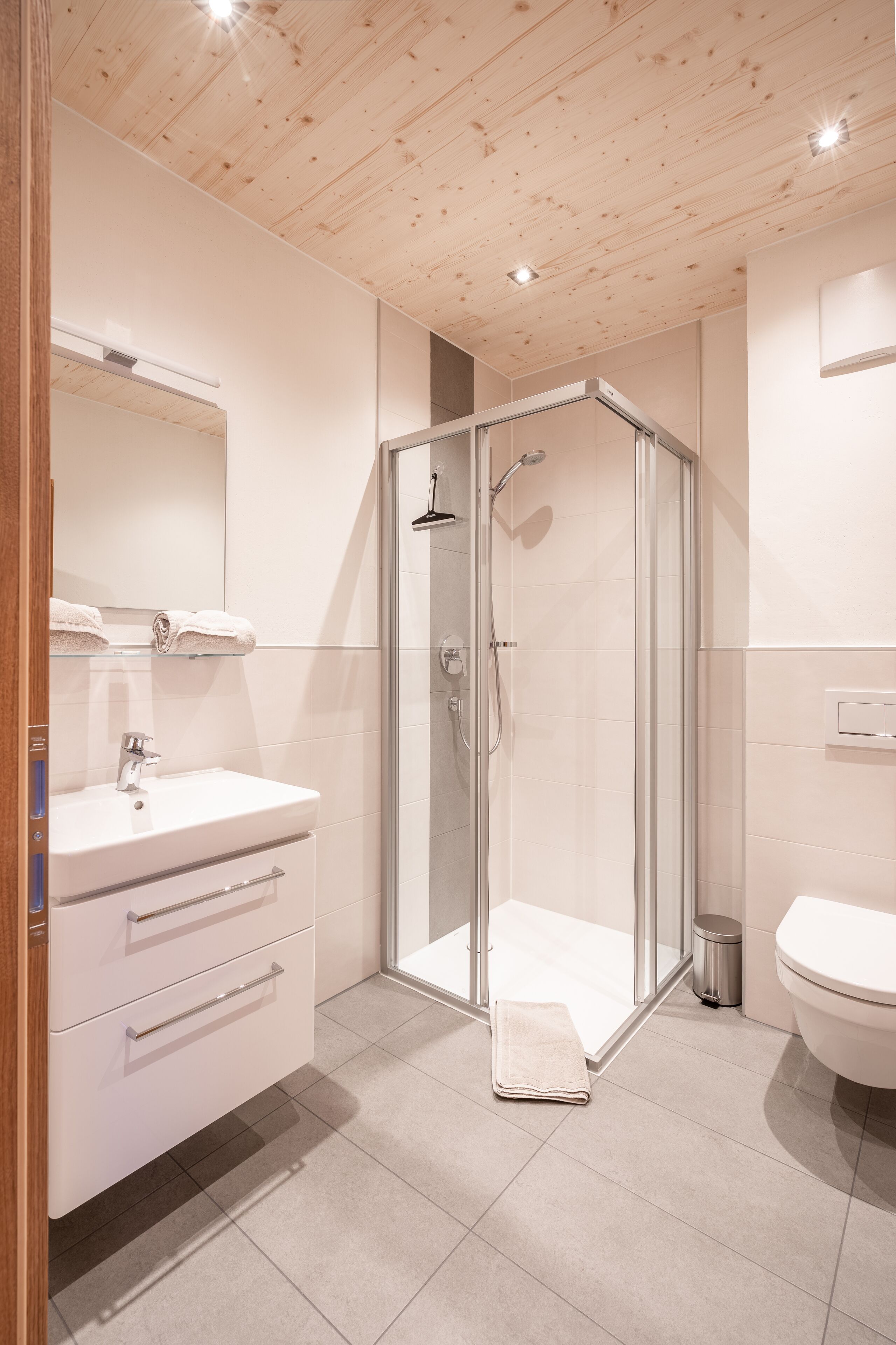Un baño moderno con techo de madera, ducha con cerramiento de vidrio, lavamanos blanco y un inodoro.