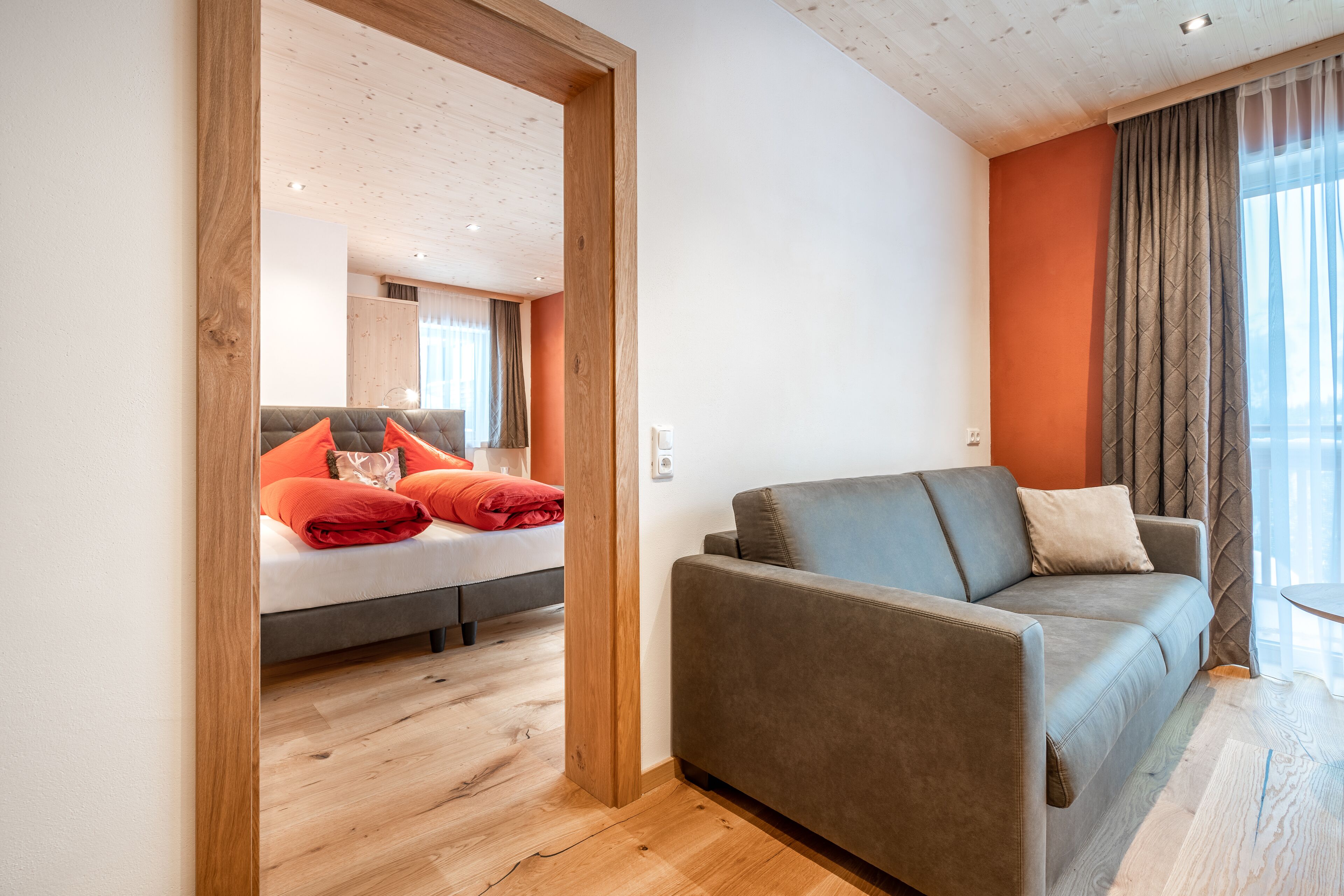 A modern living room with a grey sofa, a wooden floor, and a bed with orange pillows.
