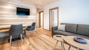 A modern living room with a wooden ceiling, a flat-screen TV mounted on the wall, a grey sofa, a coffee table, and a dining area with chairs and a table. - Naturhaus Gehren (Steeg)