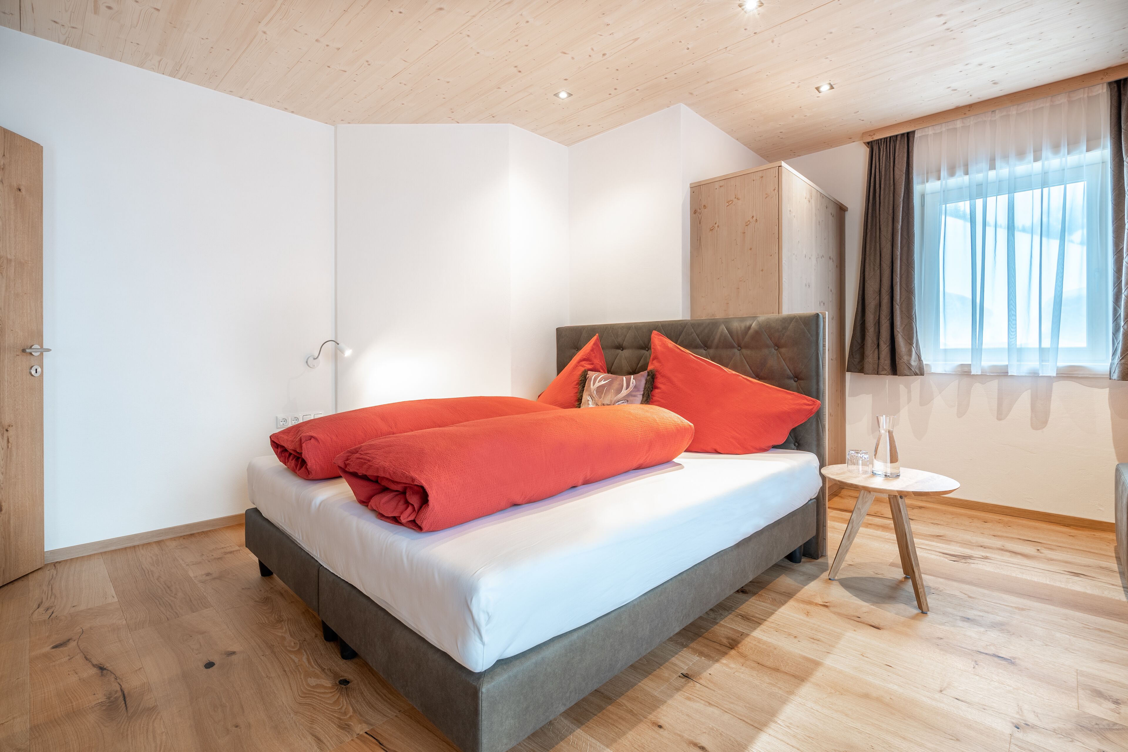 A bedroom with a bed, a bedside table, a window with curtains, and wooden flooring.
