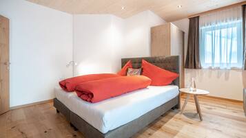 A bedroom with a bed, a bedside table, a window with curtains, and wooden flooring.