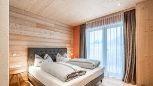 A bedroom with a wooden ceiling, two beds with grey bedding, a bedside table, and a window with curtains. - Naturhaus Gehren (Steeg)