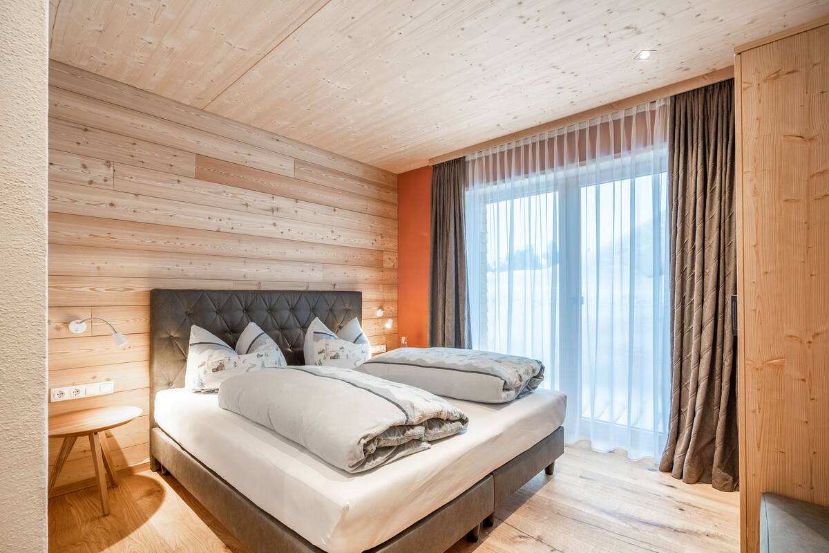 Un dormitorio con techo de madera, dos camas con ropa de cama gris, una mesita de noche y una ventana con cortinas.
