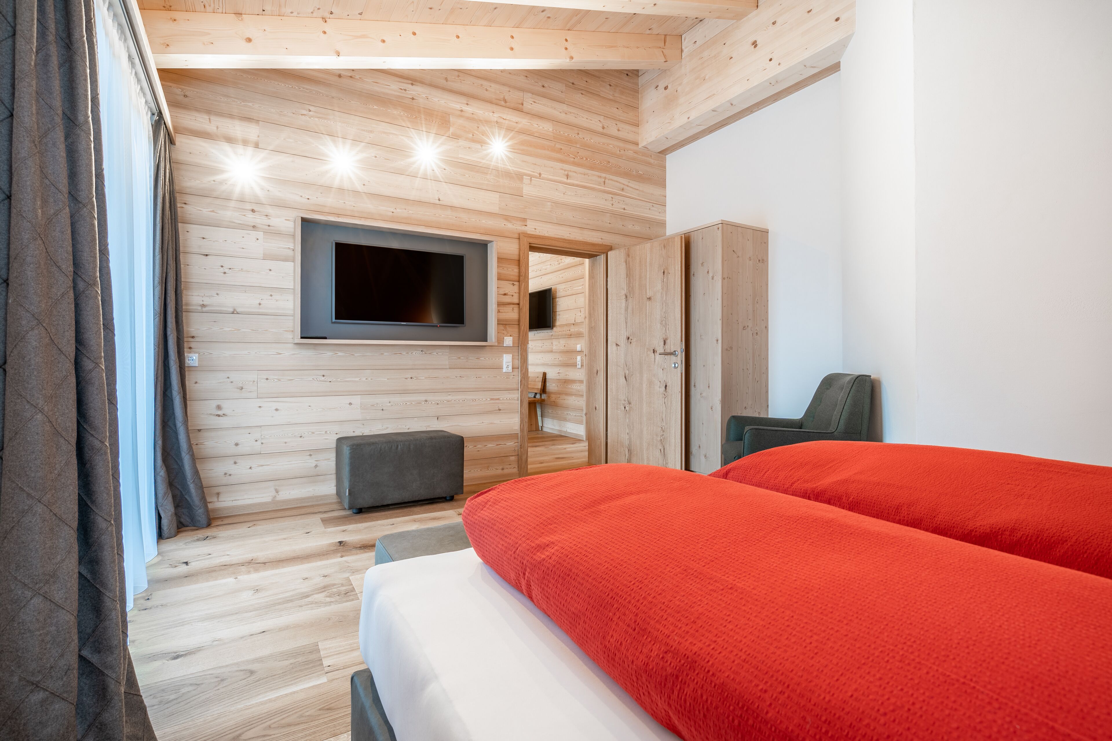 Ein modernes Zimmer mit Holzwandverkleidung, einem an der Wand montierten Flachbildfernseher, einem Bett mit roter Bettwäsche und einer grauen Bank.