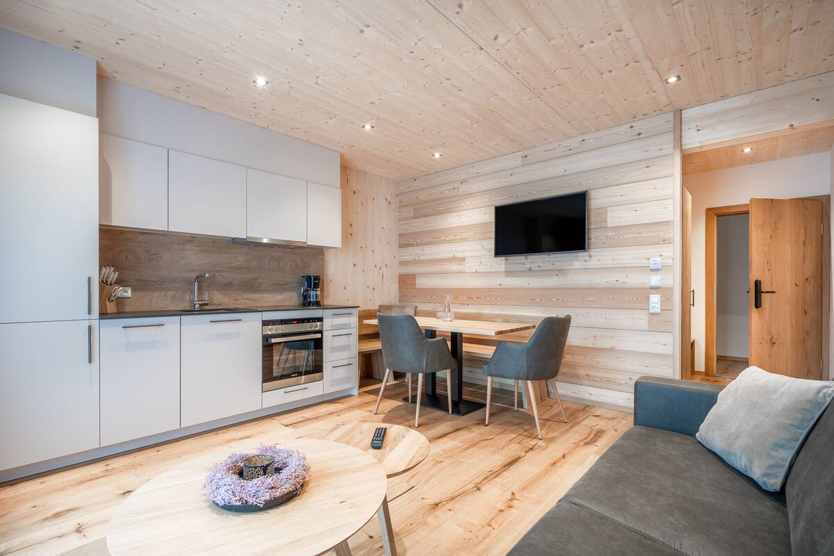 Una cocina moderna con armarios blancos, una mesa de comedor de madera con dos sillas y un televisor de pantalla plana montado en la pared.