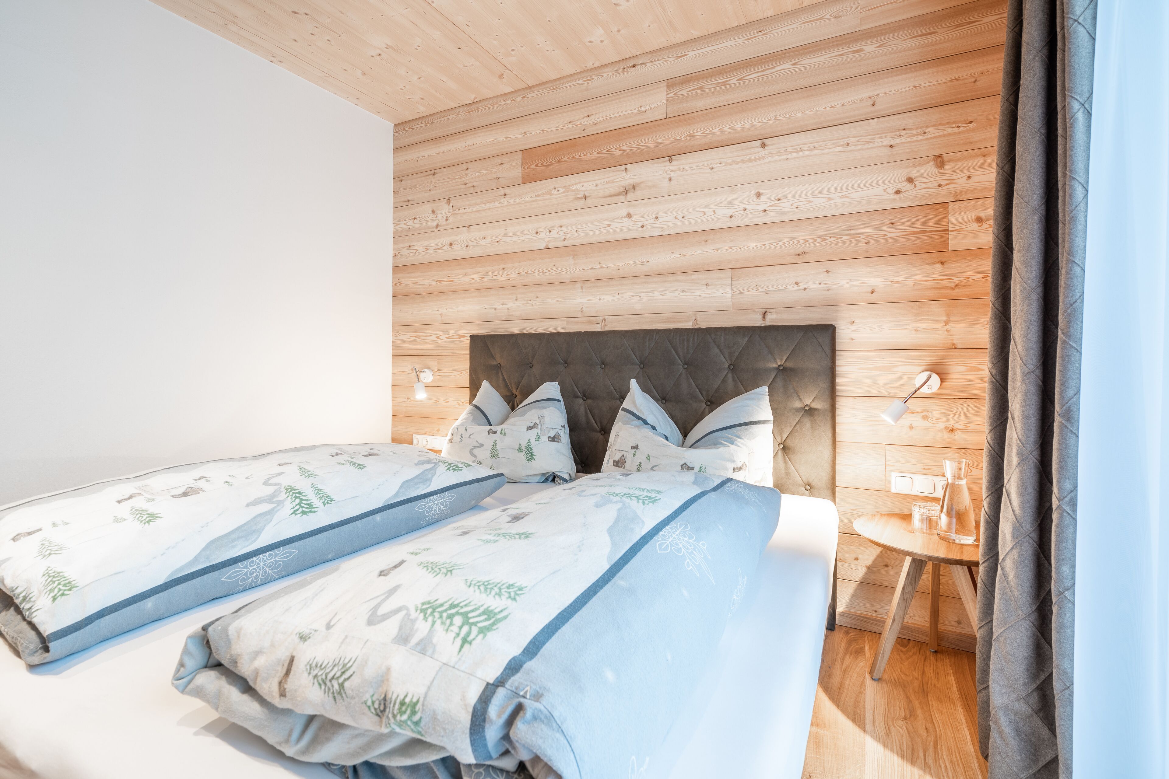 A bedroom with a wooden ceiling, a tufted headboard, two beds with matching bedding, bedside tables with lamps, and a small wooden table with a glass.
