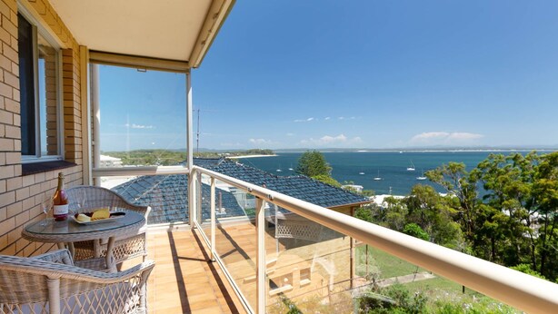 Een balkon met een tafel, stoelen en een fles champagne. Vanuit dit balkon is er uitzicht op een kustgebied met boten en een heldere lucht.