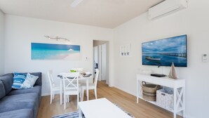 Een moderne woonkamer met een grijze bank, een witte eettafel met stoelen, een flatscreen televisie en een schilderij van een strandgezicht.
