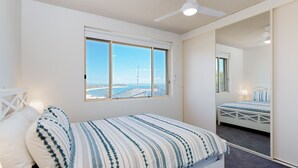 Slaapkamer met een groot raam met uitzicht op de kust, een plafondventilator en een bed met gestreept beddengoed.