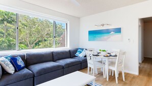Een moderne woonkamer met een grijze bank, een witte eettafel en stoelen. Een groot raam biedt uitzicht op bomen en een gebouw.