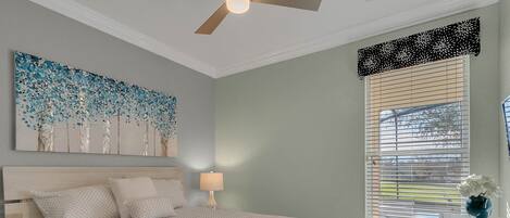 Une chambre à coucher comprenant un lit, un ventilateur de plafond, une fenêtre avec des stores, une table de chevet avec une lampe et une œuvre d’art murale.