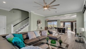 A spacious living room with a brown leather sofa, a glass coffee table, and a ceiling fan. A staircase leads to an upper level, and a dining area is visible in the background. - Disney 6BR | Theater | Themed Bedrooms | Pool/Spa (Kissimmee)