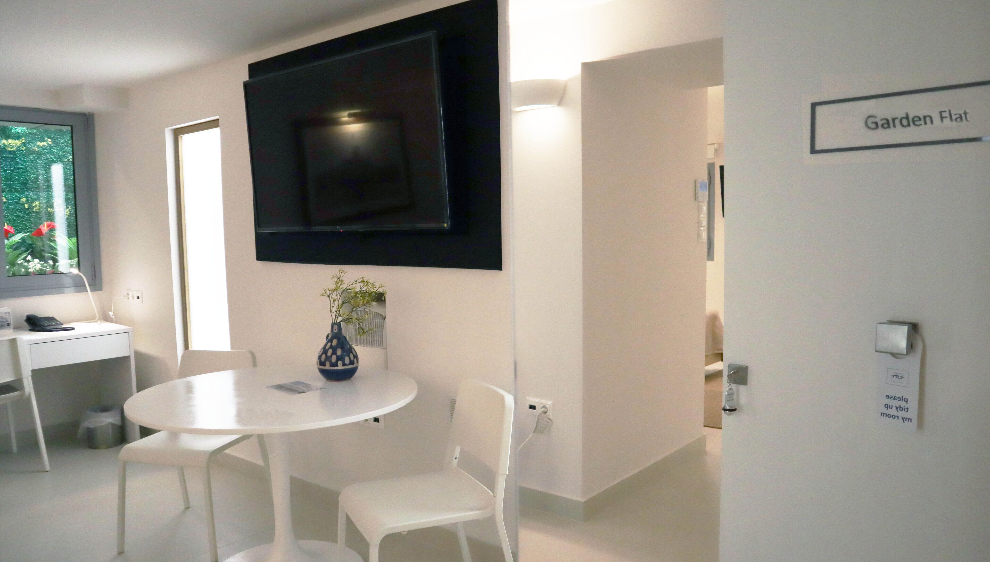 A modern hotel room with a flat-screen TV mounted on the wall, a small round table with two white chairs, a vase with a plant, and a sign indicating the room is on the Garden Flat.
