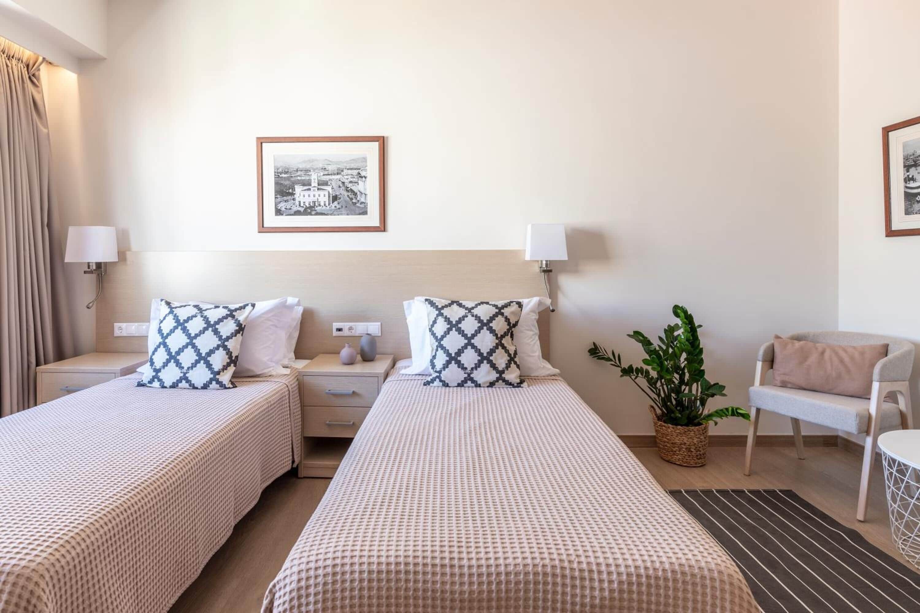 Habitación de hotel con dos camas, una mesita de noche, una lámpara, una silla y un cuadro en la pared.