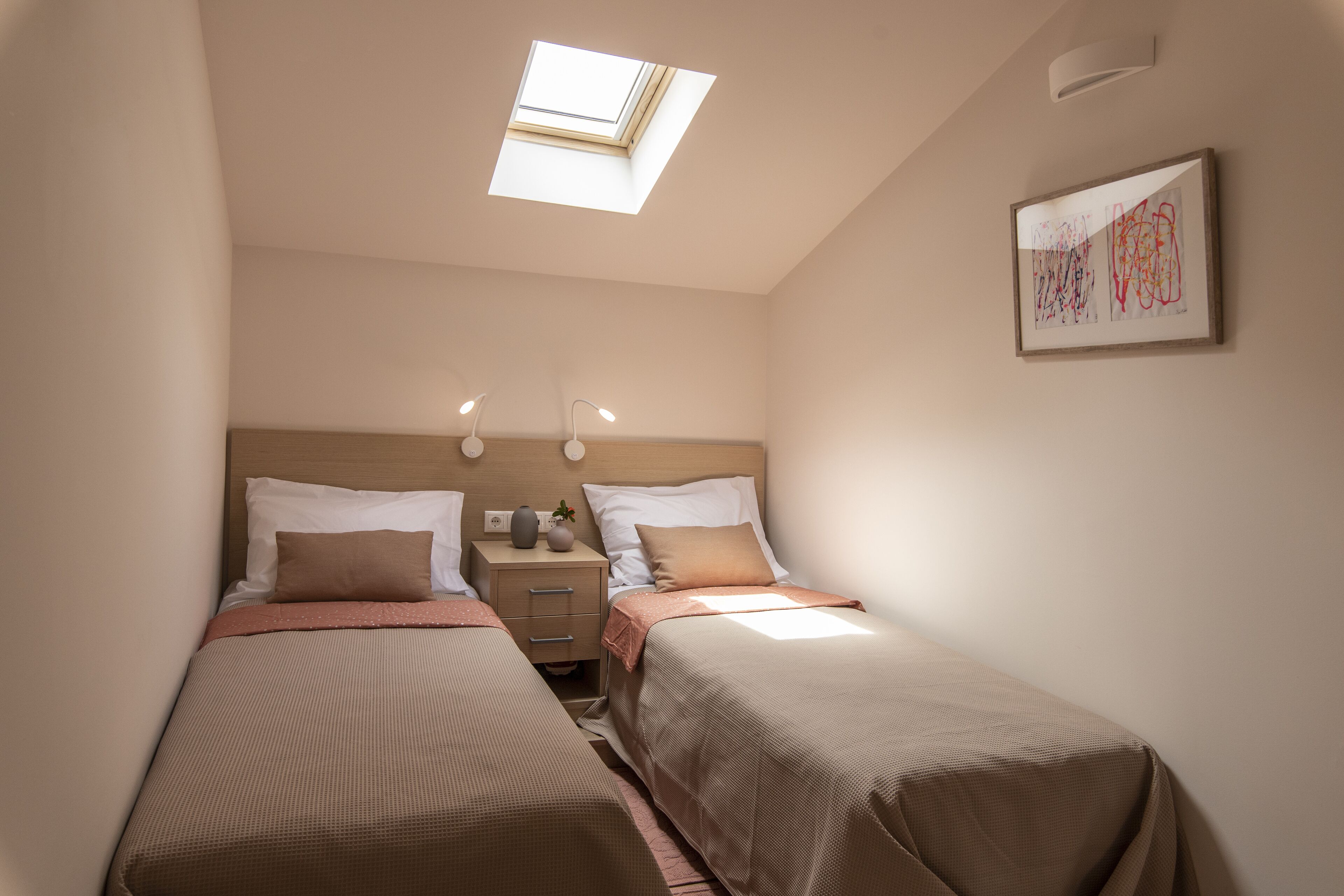 A bedroom with two beds, a skylight, bedside lamps, a nightstand, and a framed abstract painting.