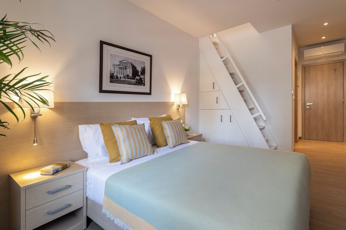 Un dormitorio con cama, mesitas de noche, una lámpara, un cuadro en la pared y una escalera.