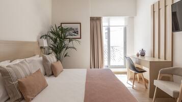 Una habitació d'hotel amb llit, escriptori, cadira, planta en test i finestra amb cortines.
