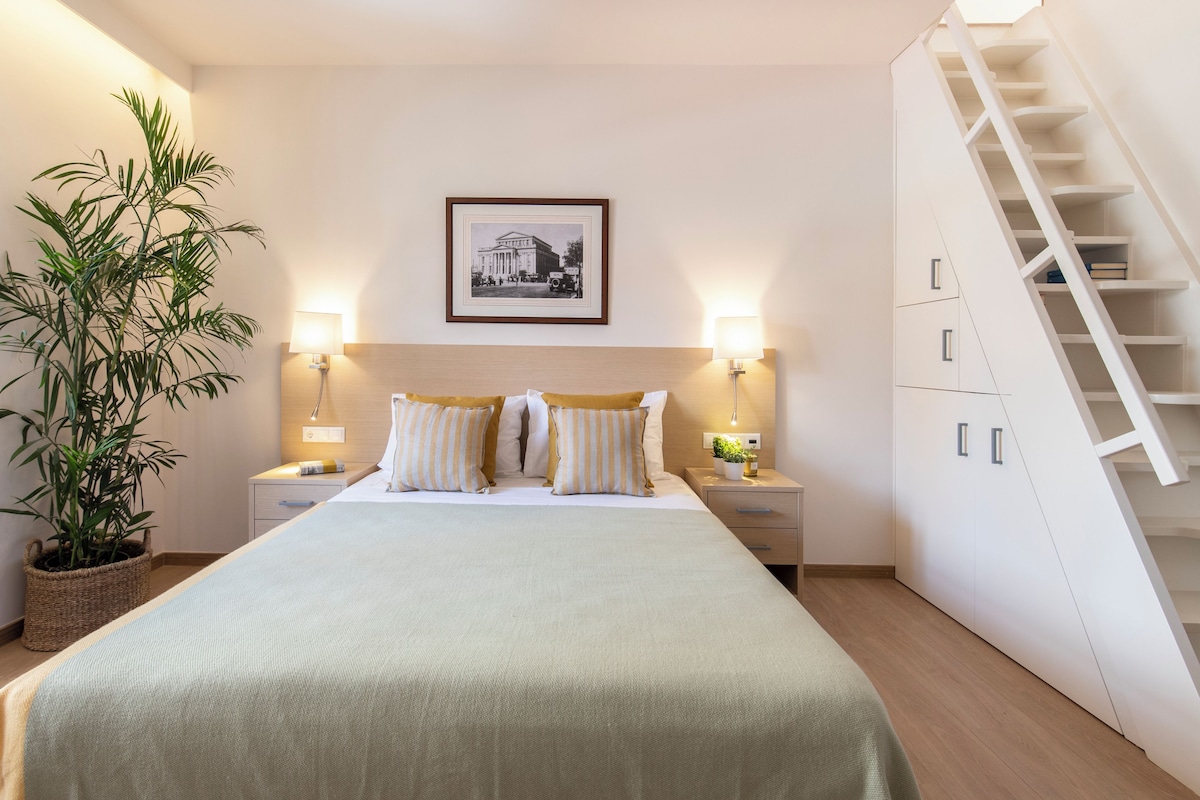 A neatly arranged bedroom with a bed, bedside tables, a potted plant, a framed picture on the wall, and a staircase.
