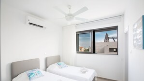 Ein Hotelzimmer mit zwei Betten, einem Deckenventilator, einer Klimaanlage, einem großen Fenster mit Blick auf Gebäude und einem Gemälde an der Wand.