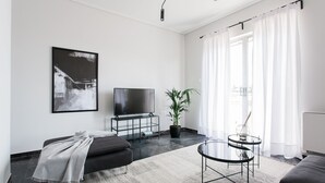 A modern living room with a flat-screen TV, a sofa, a coffee table, a potted plant, and a large window with sheer curtains. - Spacious Apartment near HELEXPO (Marousi)