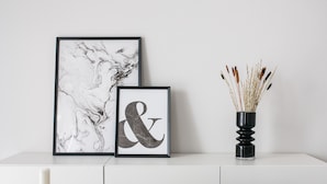 A white console table with two framed artworks, a black vase with dried reeds, and a green upholstered chair. - Spacious Apartment near HELEXPO (Marousi)