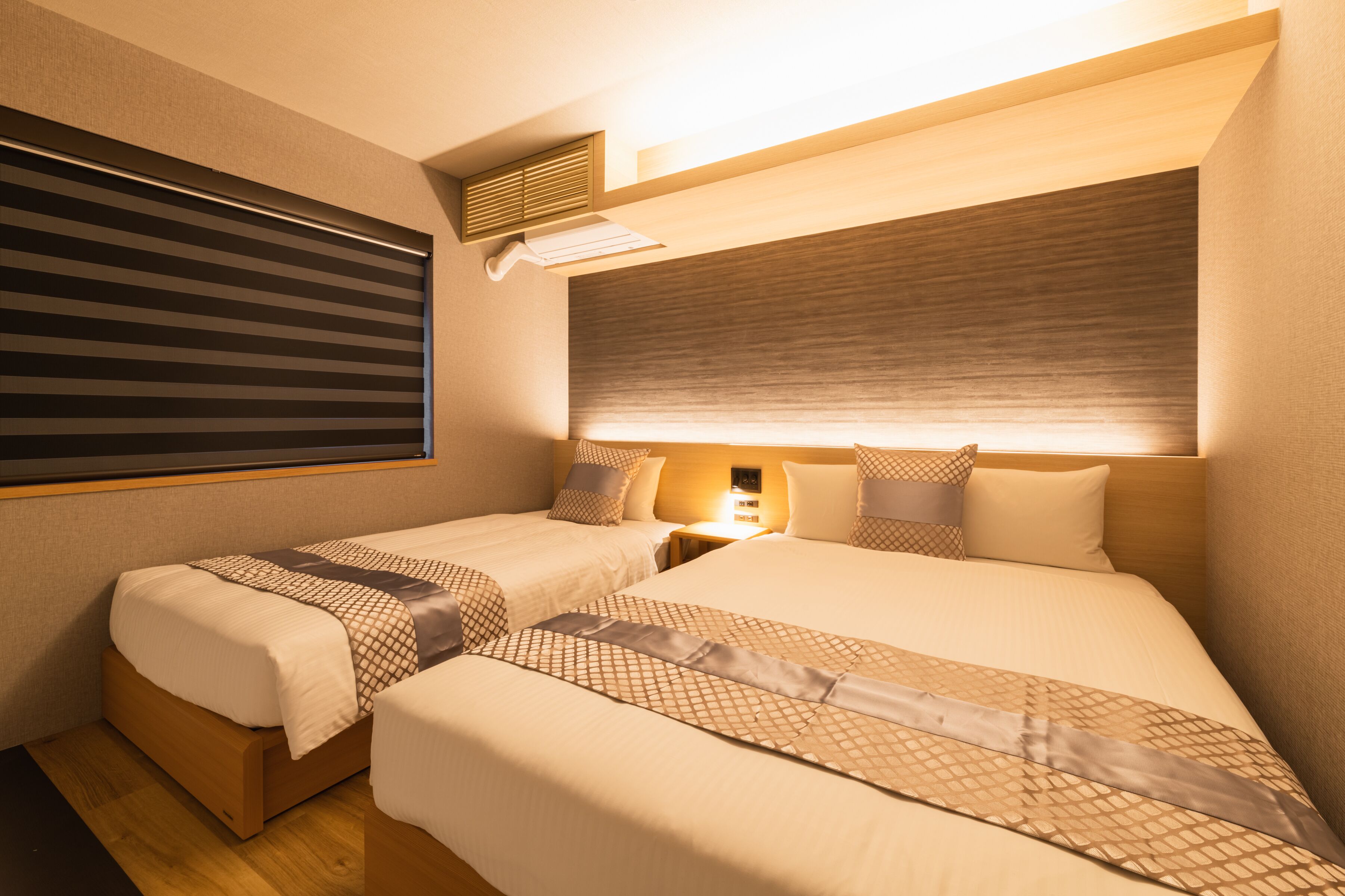 Une chambre d’hôtel avec deux lits, une grande fenêtre avec stores, une table de chevet avec une lampe et une bouche d’aération fixée au mur.