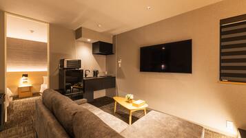 A modern living room with a sofa, a small table with a vase, a flat-screen TV mounted on the wall, and a kitchen area with a microwave and a coffee maker.