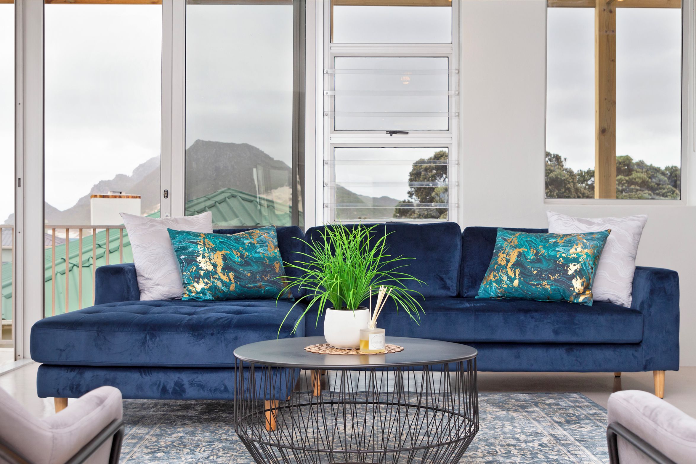 A modern living room with a dark blue sofa, white and teal patterned cushions, a round coffee table with a black wireframe base, and a potted plant.