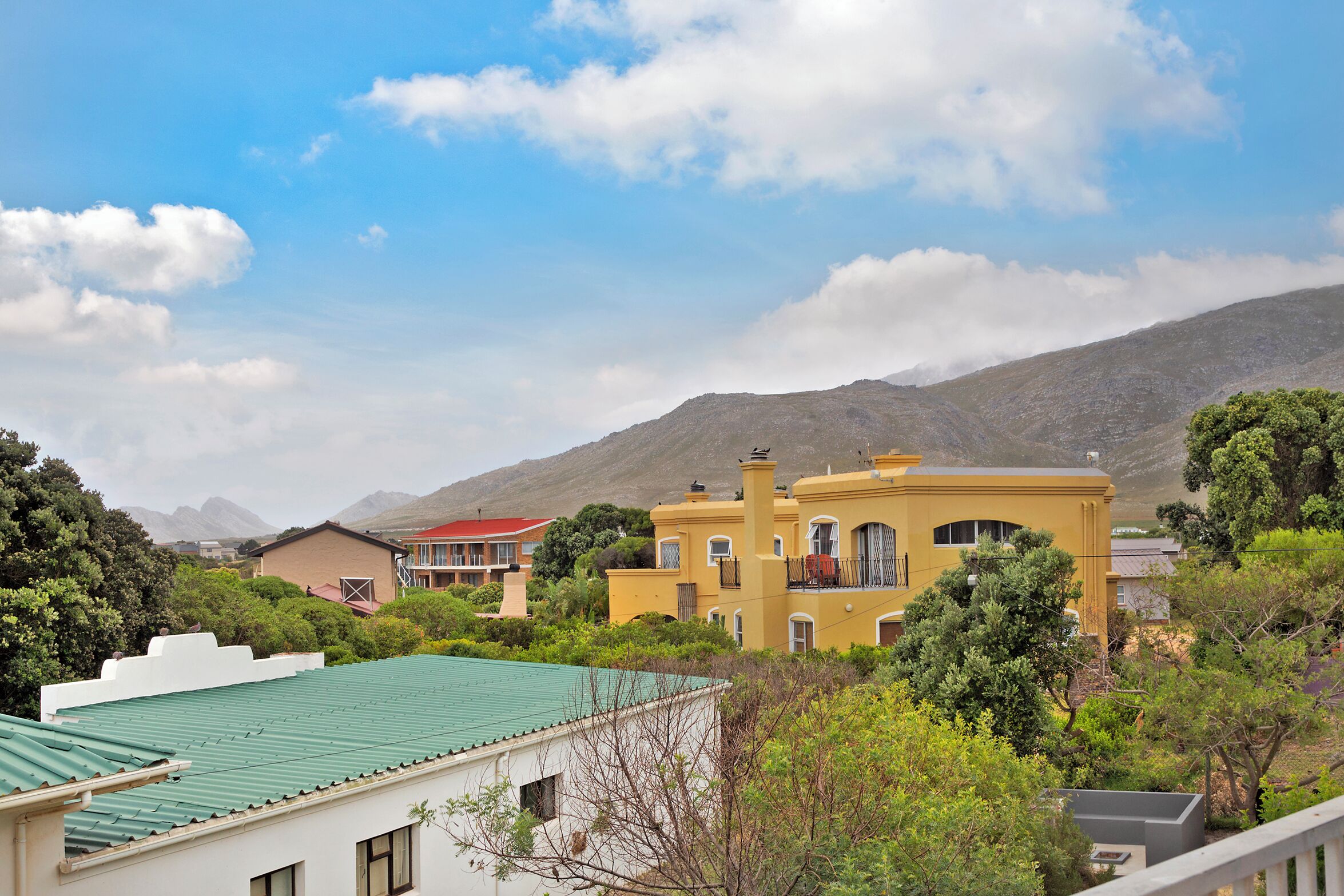 A yellow building with a green roof, surrounded by trees and other buildings, with mountains in the background.