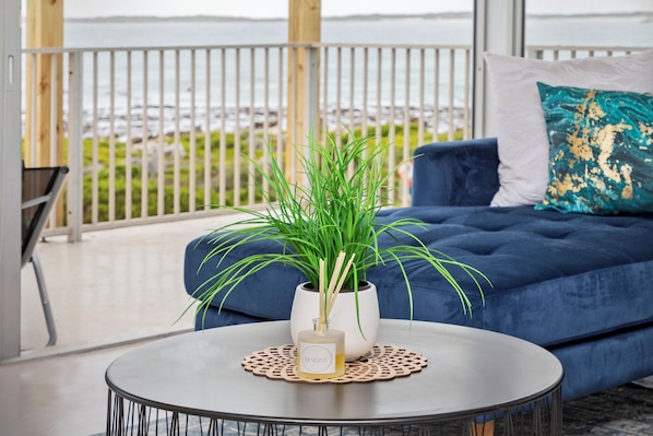 A balcony with a view of the sea, a blue sofa, a white vase with green plants, and a round table with a bottle of perfume.