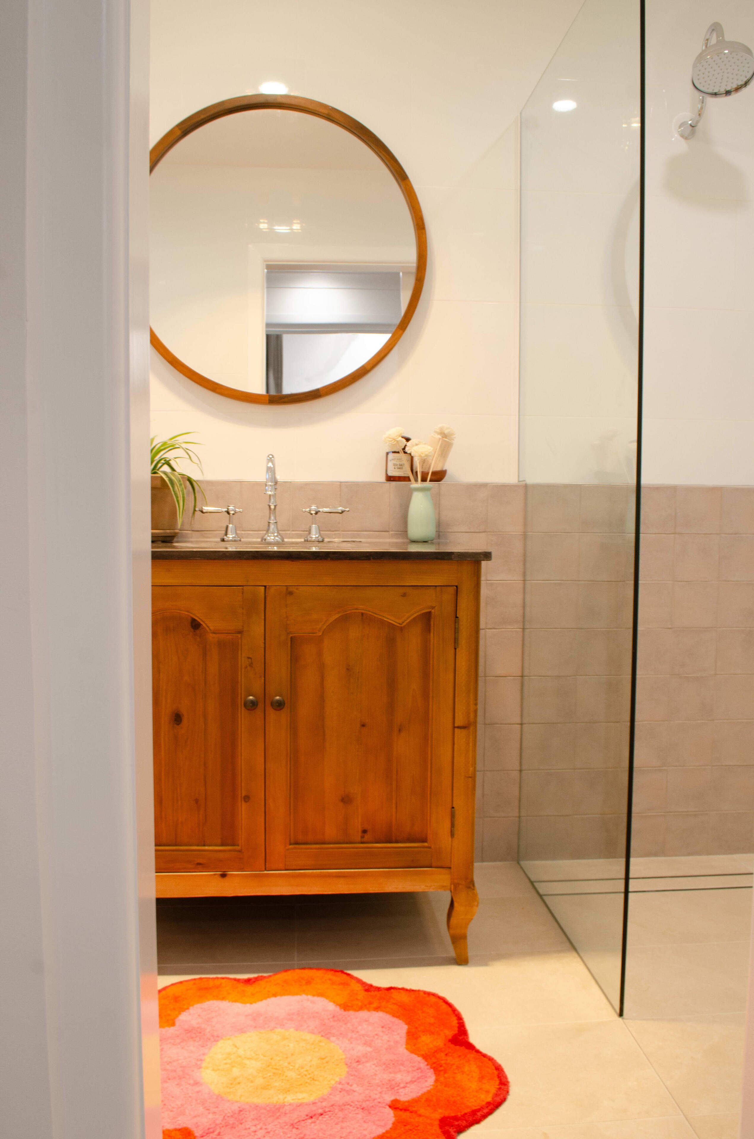 A bathroom with a wooden vanity, a round mirror, a shower, and a pink floral rug.