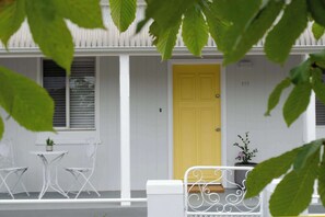 A white building with a yellow door, a white porch with two chairs and a table, and a small potted plant.