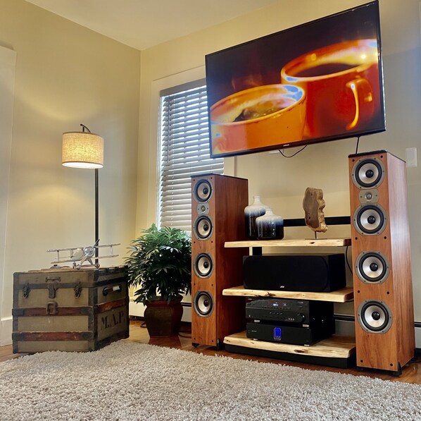 Una sala de estar con un mueble de madera para centro de entretenimiento, un televisor de pantalla plana, una lámpara, una planta en maceta y una maleta antigua.