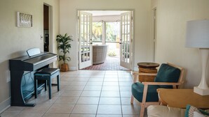 A room with a piano, a bench, a chair, a lamp, and a view through an open door to a balcony. - Tiki Retreat (North Fort Myers)