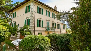A two-story building with green shutters, a balcony with a wrought iron railing, and a garden area with a lawn and bushes.