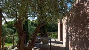 Un terrazzo in legno con tavolo e sedie, circondato da alberi e un edificio.